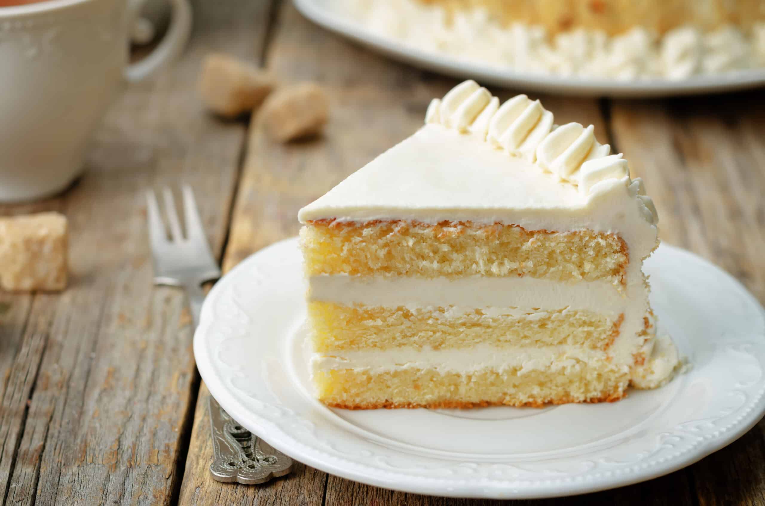 Slice of three-layered sponge cake with buttercream icing on a small white plate with dainty fork on the left, photographed on a rustic wooden table